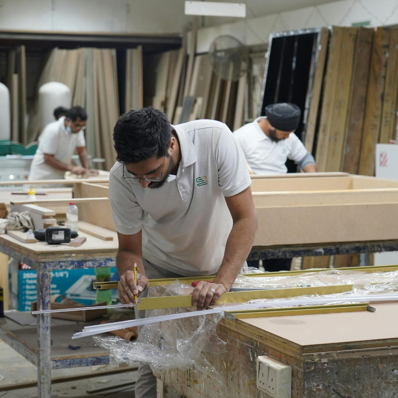 Man measuring wood in a workshop with other workers in the background