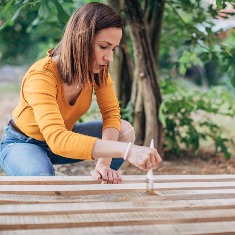 Woman painting wooden slats outdoors with greenery in the background