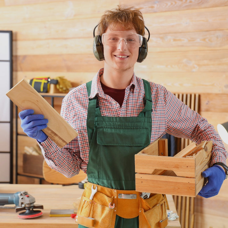 Man in a workshop holding wooden pieces, wearing safety goggles and ear protection.